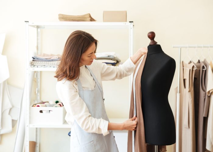 Brunette seamstress in apron measuring beautiful fabric on black dummy in workshop